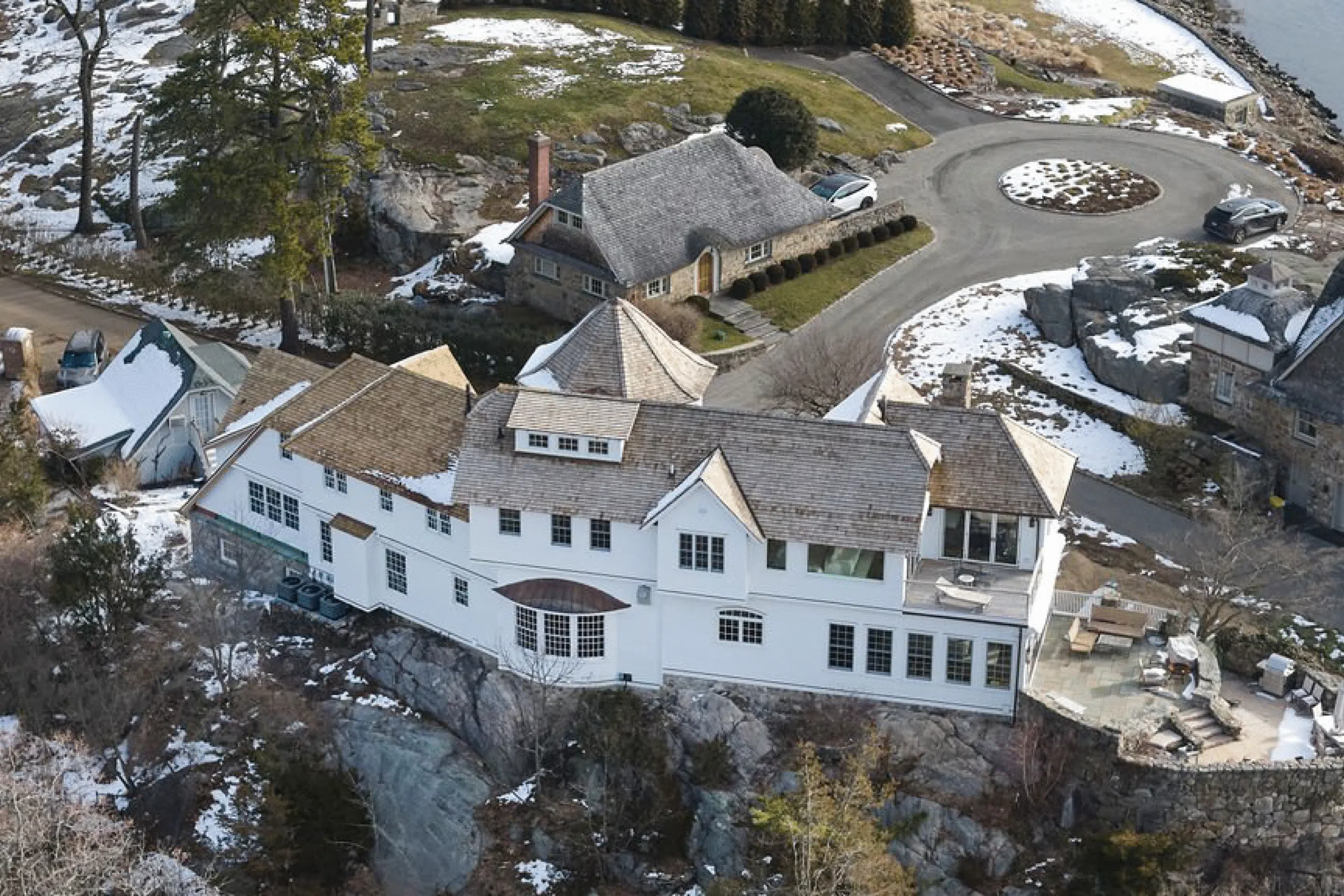 Aerial view of a large white house with wooden roofing built on a rocky cliff, surrounded by snow